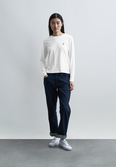 Γυναίκα που στέκεται με τα χέρια εν μέρει στις τσέπες, φορώντας λευκό μακρυμάνικο πουκάμισο, σκούρο τζιν με ρεβέρ και λευκά αθλητικά παπούτσια, μπροστά σε μονόχρωμο φόντο.