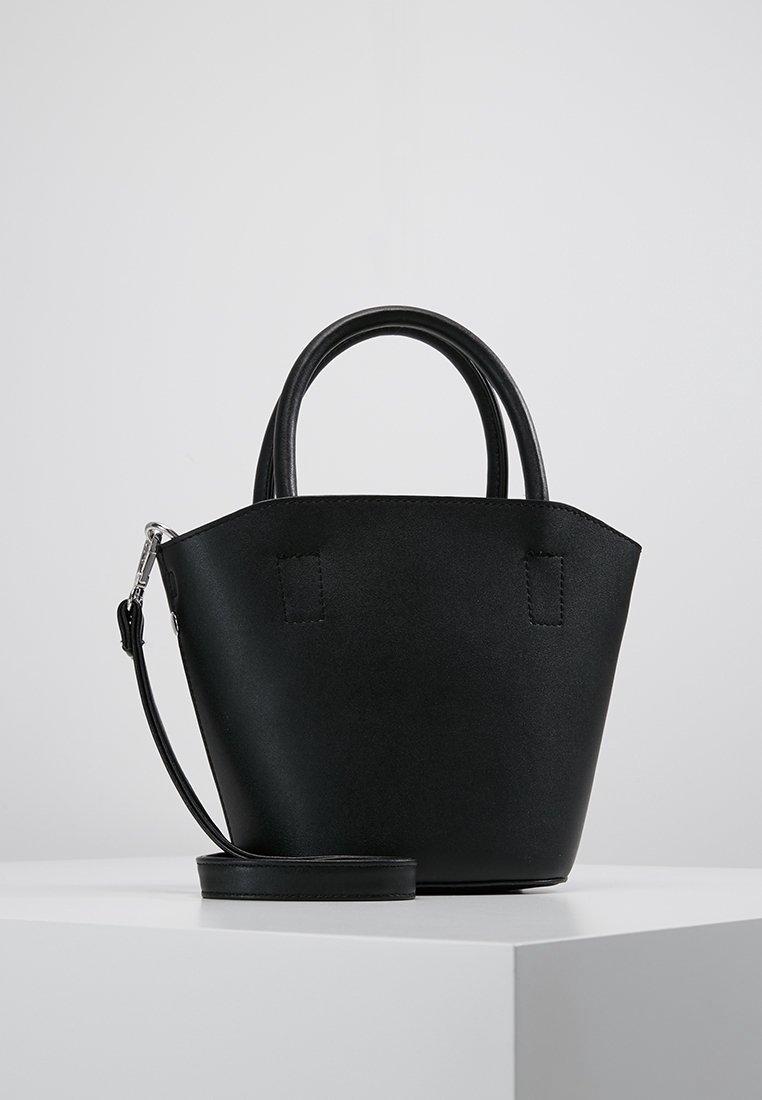 Sac à main en cuir noir avec deux poignées courtes et une longue bandoulière amovible, posé sur une surface blanche contre un fond clair uni.