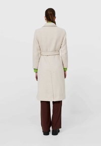 Manteau long de couleur crème avec une taille ceinturée et un col en notch. Présente des accents de poignets verts. Coupe droite, tissu texturé et longueur mi-saison.