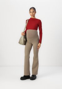 Zwangerschapsoutfit met een rode geribde top met lange mouwen en beige flared broeken met een ondersteunende tailleband. Zwarte platform schoenen en een groene tas.