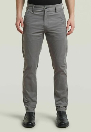 Homme portant un pantalon gris ajusté et des chaussures en cuir noires, debout devant un fond clair uni.
