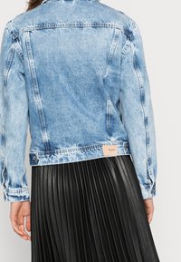 Light blue denim jacket with faded texture, button cuffs, and a branded label on the back hem, paired with a black pleated skirt.