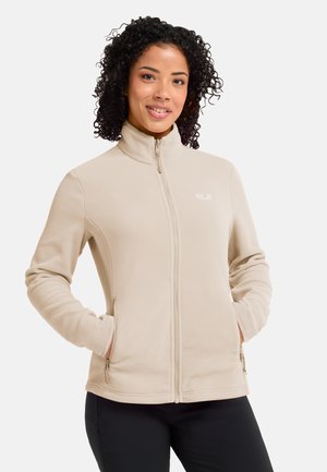 Frau trägt eine beige Fleecejacke mit durchgehendem Reißverschluss, Hände in den Seitentaschen, lächelnd und steht vor einem schlichten hellen Hintergrund.