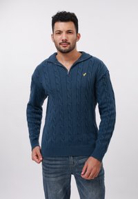 Bărbat cu păr creț scurt și barbă, îmbrăcat cu un pulover albastru cu model tricotat, cu fermoar până la jumătate, și blugi albaștri, stând în fața unui fundal alb.