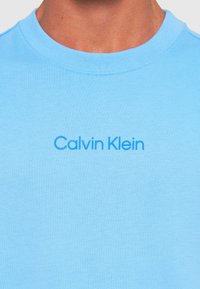 Bărbat îmbrăcat cu un tricou de culoare bleu deschis, cu guler rotund, pe care este imprimat în albastru logo-ul "Calvin Klein" pe piept.