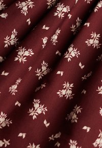 Vinrødt stof med et delikat hvidt blomstermønster, der indeholder små blomster og blade. Glat tekstur med en let draperet fremtoning.