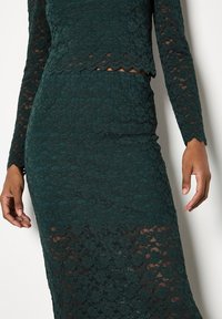 Vestido verde de encaje con mangas largas, silueta ajustada, dobladillo con forma de ondas y patrones florales en toda la tela texturizada.