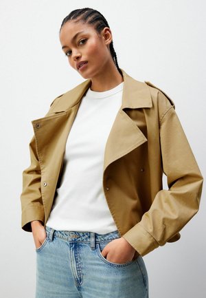 Beige leichte Jacke mit doppelter Knopfleiste, großem Kragen und seitlichen Taschen, getragen über einem weißen Rundhals-Shirt und blauen Jeans.