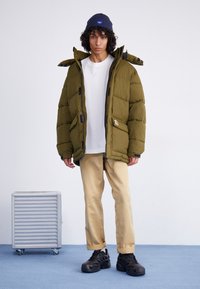 Oliwkowa puchowa kurtka z zamkiem z przodu i dużym kapturem, w połączeniu z białą koszulą i beżowymi spodniami. Czarne masywne buty i niebieska czapka beanie.