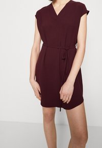 Robe courte bordeaux avec un décolleté en V, des manches courtes et une ceinture à nouer à la taille. Le tissu a une finition texturée.