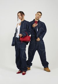 Dva modeli nosita usklajene temne denim jakne in hlače z svetlimi šivi. Ena drži rdečo torbico, druga pa rdeč telefon.