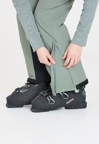 Skihosen aus hellgrünem Stoff mit Reißverschlüssen an den Säumen, schwarzen Akzenten und glatter Textur, kombiniert mit schwarzen Skischuhen.
