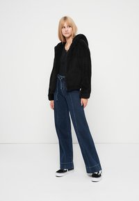 Μαύρο, χνουδωτό hoodie με εμπρόσθιο φερμουάρ και κουκούλα, σε συνδυασμό με ψηλόμεσα τζιν παντελόνια φαρδιάς γραμμής. Τα παπούτσια είναι μαύρα και λευκά αθλητικά παπούτσια.