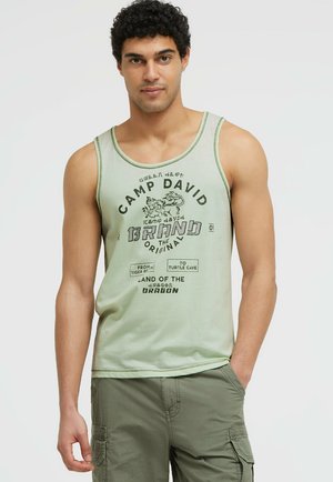 Mann trägt ein hellgrünes Camp David Tanktop und grüne Cargo-Shorts, steht mit einer Hand in der Tasche vor schlichtem Hintergrund.