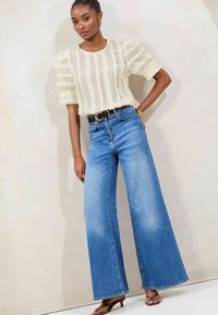 Light yellow blouse with ruffled sleeves, high-waisted blue wide-leg jeans, and brown belt, paired with brown heeled sandals.