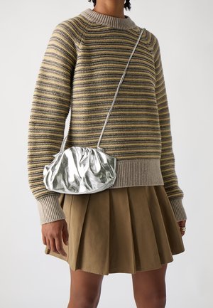 Personne portant un pull en maille rayé et une jupe plissée marron, avec un petit sac à bandoulière argenté et froncé.