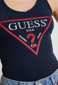 Marineblauwe geribbelde body met een rood driehoekig logo met "GUESS U.S.A." en een vraagteken, gecombineerd met lichtblauwe denim shortss.