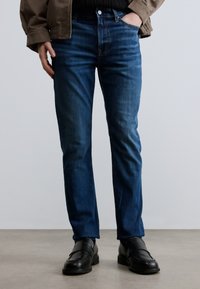 Σκούρο μπλε τζιν με ίσια γραμμή, με κλασικό σχέδιο πέντε τσεπών, υφή από τζιν και διακριτικό εφέ ξεθωριάσματος. Συνδυάζονται με μαύρα παπούτσια.