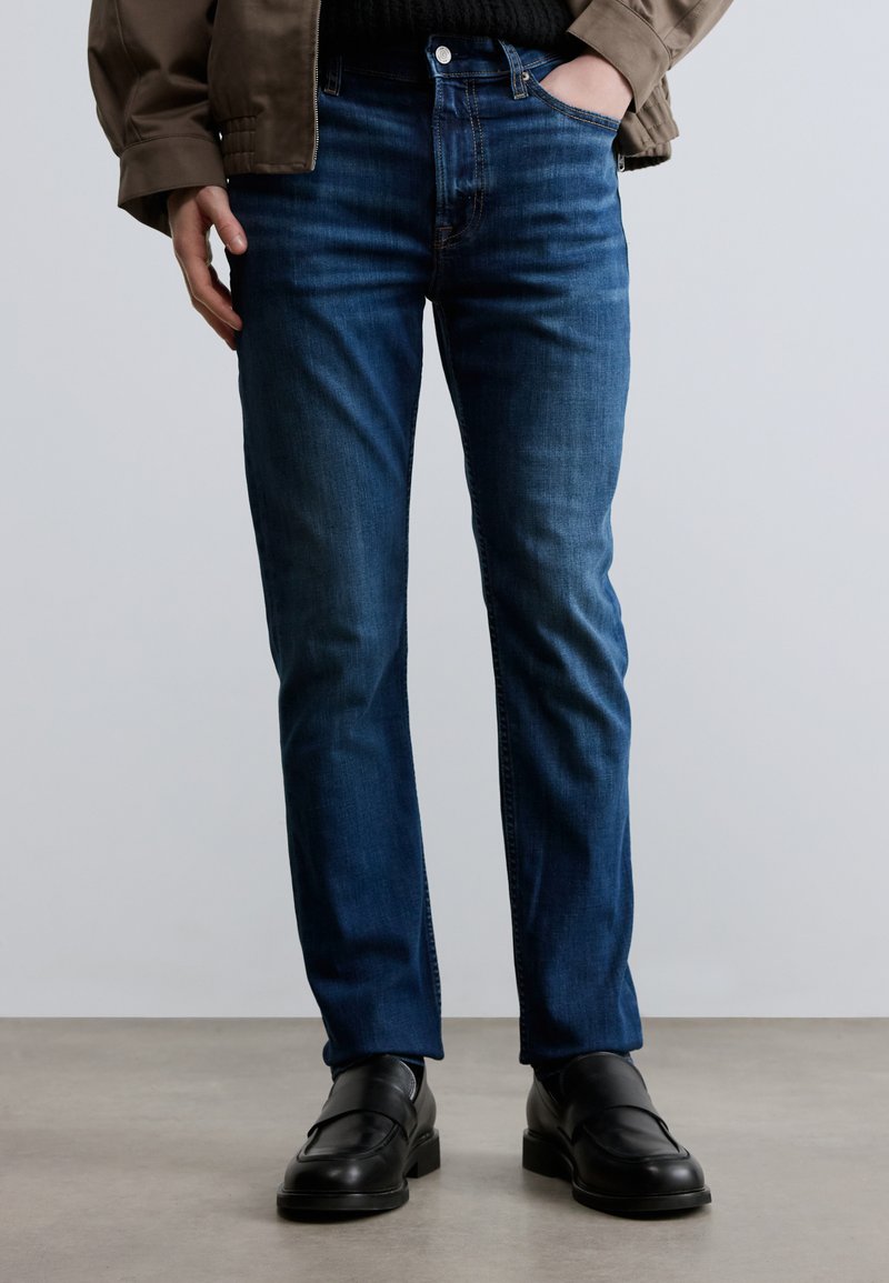 Σκούρο μπλε τζιν με ίσια γραμμή, με κλασικό σχέδιο πέντε τσεπών, υφή από τζιν και διακριτικό εφέ ξεθωριάσματος. Συνδυάζονται με μαύρα παπούτσια.