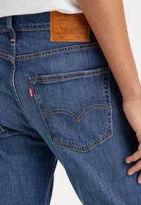 Gros plan sur une personne portant un jean Levi's bleu avec un patch de marque en cuir visible, des coutures sur la poche arrière et une étiquette rouge.