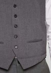 Zbliżenie na osobę w szarej kamizelce zapinanej na guziki, noszącą białą koszulę, z widocznymi w dolnej części szarymi spodniami w wzory.