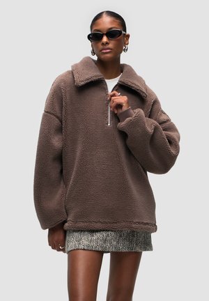 Brauner Fleece-Pullover mit hohem Kragen, Viertelreißverschluss vorne und lockerer Passform. Mit einer weichen Textur und gerippten Bündchen. Tragen über einem gemusterten Rock.