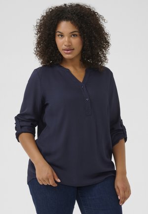 Blusa blu navy con bottoni, scollo a V, maniche arrotolabili e tessuto morbido. Presenta una leggera lucentezza e una vestibilità aderente. Abbinata a jeans scuri.