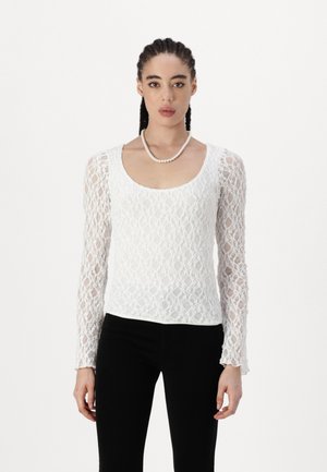 White lace long-sleeve top with a scoop neckline, featuring intricate patterns and textured fabric. Paired with black pants.
