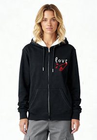 Sweat à capuche noir zippé avec doublure en polaire blanche et deux poches avant. Présente le mot "amour" en blanc avec un accent rouge. Manches longues avec poignets côtelés.