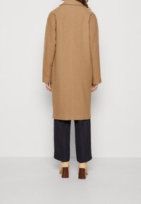 Frakke i kameluld blanding med en lige silhuet, bagventilation og brede ærmer. Kombineret med mørkeblå koniske bukser og beige hæle.