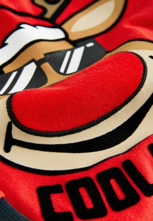 Punane kangas, millel on naeratava põhjapõdra graafika, kes kannab päikeseprille. Kujundus sisaldab pehmet plush-tekstuuri ja musti ääriseid.