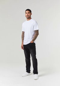 Λευκό βαμβακερό t-shirt σε συνδυασμό με μαύρο τζιν και λευκά αθλητικά παπούτσια. Το μοντέλο στέκεται με αυτοπεποίθηση, παρουσιάζοντας ένα casual σύνολο με εφαρμοστή γραμμή.