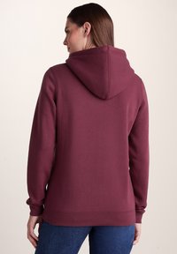 Woman wearing a maroon hooded sweatshirt viewed from the back, paired with blue jeans against a plain light background.