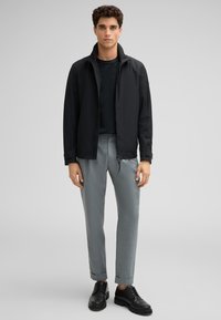 Giacca nera zip-up con colletto alto, abbinata a un top scuro aderente e pantaloni grigi chiari a gamba stretta. Le scarpe nere stringate completano il look.