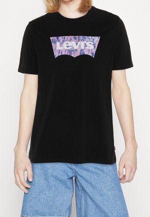 Person iført en sort Levi's t-shirt med et pink og lilla logo, kombineret med lyseblå denimshorts mod en hvid baggrund.