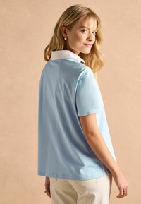 Svetlo modrá polokošeľa s krátkym rukávom, vybavená bielym golierom, voľným strihom a bočnými rozparkmi pre pohodlný pohyb. Hladký bavlnený materiál.