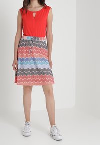 Top rojo sin mangas con un corte en forma de llave, combinado con una falda de patrón ondulado multicolor en azul, rojo y negro; zapatillas blancas.