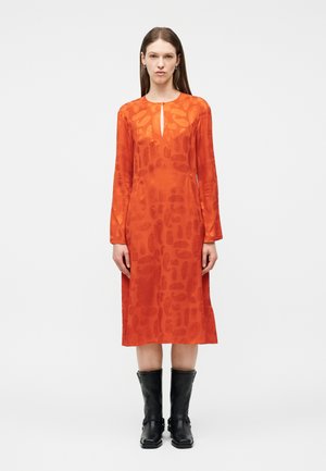 Γυναίκα που φοράει πορτοκαλί midi φόρεμα με μακριά μανίκια και διακριτικά σχέδια, σε συνδυασμό με μαύρες δερμάτινες μπότες μέσης γάμπας, στεκόμενη μπροστά σε λευκό φόντο.