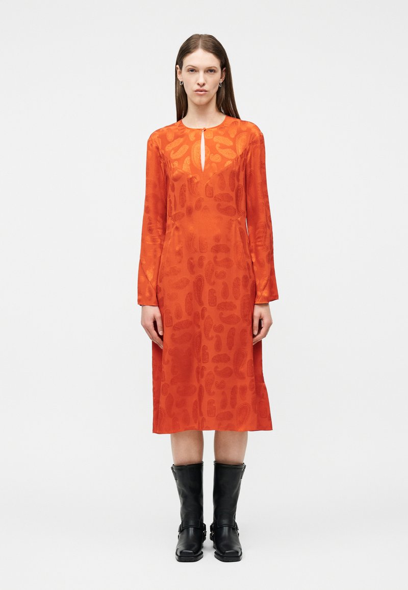 Γυναίκα που φοράει πορτοκαλί midi φόρεμα με μακριά μανίκια και διακριτικά σχέδια, σε συνδυασμό με μαύρες δερμάτινες μπότες μέσης γάμπας, στεκόμενη μπροστά σε λευκό φόντο.