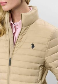 Bēšas pūkains jakas ar horizontālu quilted rakstu, augstu apkakli, priekšējo rāvējslēdzēju un mazu tumšu logo kreisajā krūtīs. Gaiši rozā krekliņš zem tā.