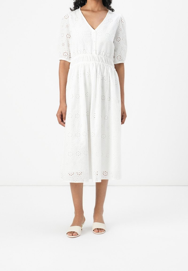 Femme portant une robe midi blanche avec motif floral à œillets, taille élastique, manches bouffantes et sandales blanches à talons ouvertes.