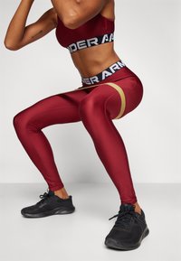 Bordo tajice s zlatnom akcentnom trakom, visokim strukom i crnim "UNDER ARMOUR" logom, glatke teksture, uparene s crnim sportskim cipelama.