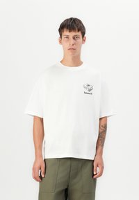 White cotton t-shirt with short sleeves, round neckline, and a small black graphic on the chest. Paired with olive green pants.