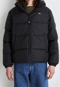 Doudoune noire avec fermeture éclair et poignets élastiques. Le design présente des panneaux matelassés horizontaux et une capuche.