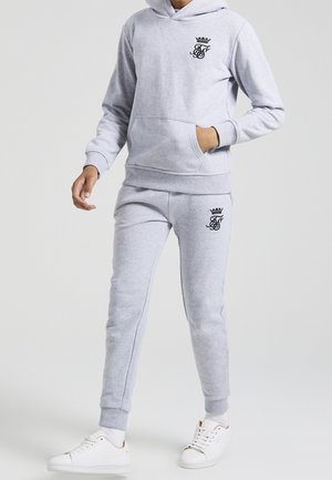 Personne portant un sweat à capuche gris clair et un pantalon de survêtement avec une couronne noire et un logo en écriture, assorti à des baskets blanches.