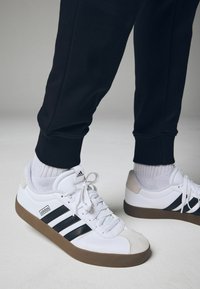 Weiße Sportschuhe mit schwarzen Streifen und grauen Akzenten, ausgestattet mit einer strukturierten Außensohle, Schnürsenkeln und gerippten schwarzen Jogginghosen.