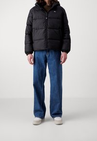 Doudoune noire avec coutures horizontales, fermeture à glissière et poignets élastiques, associée à un jean large bleu clair et des chaussures blanches.