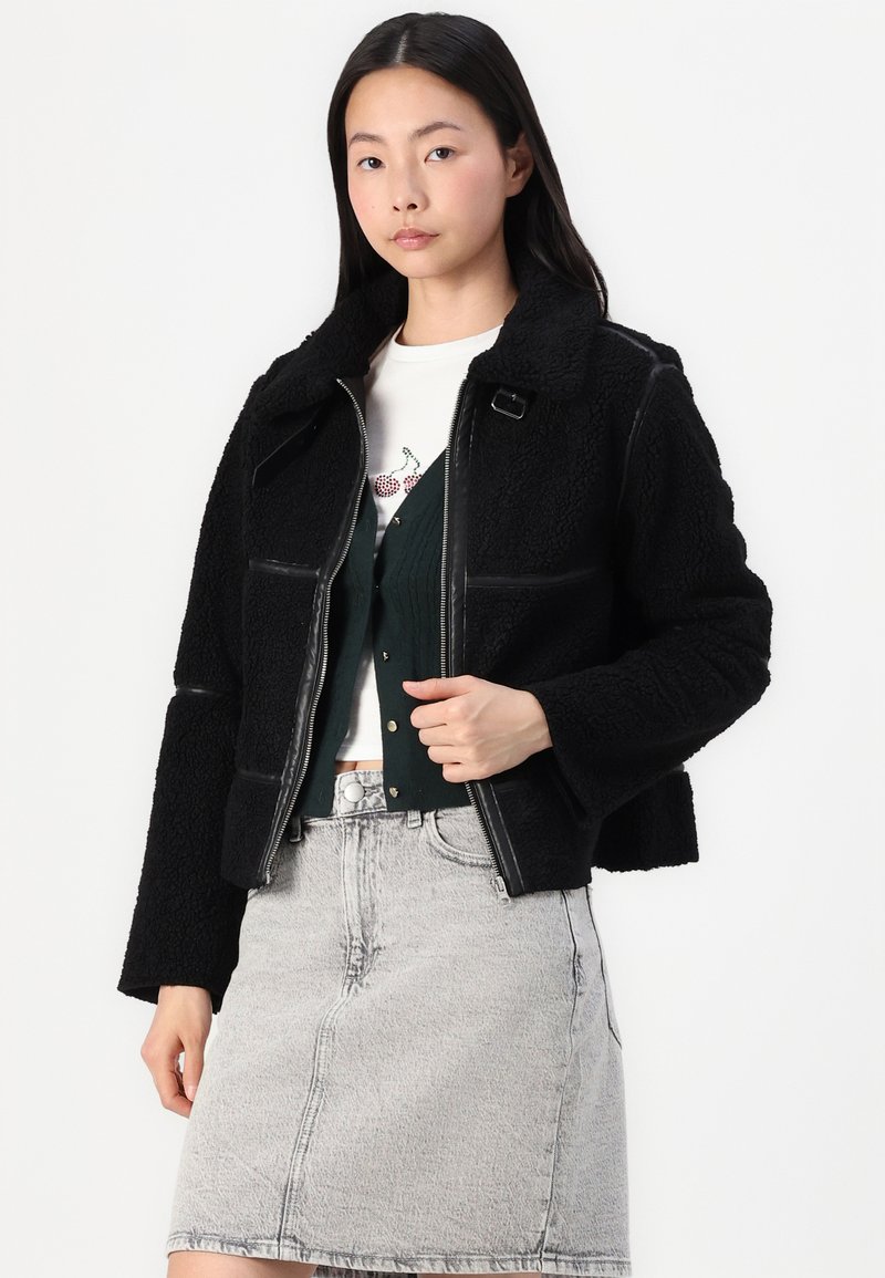 Giacca sherpa nera con dettagli in pelle, design corto, chiusura con zip e ampio collare, indossata sopra un cardigan verde e una gonna di jeans grigia.