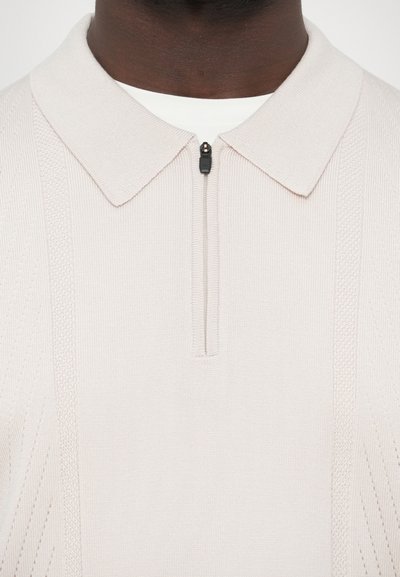 Ανοιχτό μπεζ πόλο πουκάμισο με φερμουάρ, υφασμάτινα μοτίβα και κλασικό γιακά. Κατασκευασμένο από πλεκτό ύφασμα με κάθετες ανάγλυφες λεπτομέρειες.