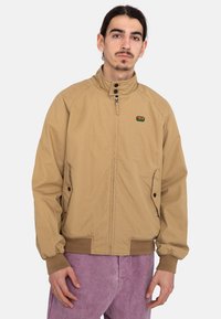 Tan jacket with a high collar, zippered front, and side pockets. Features ribbed cuffs and a small multi-colored logo on the chest.